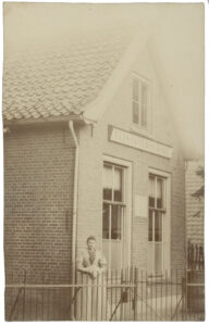 Foto van een klein gebouwtje. Aan de kopse kant twee ramen breed en onder het zadeldag één raam. Onder het bovenste raam hangt een bord "Elisabeth-school". Om de school zit een hek, achter het hek staat een vrouw in een 19e eeuwse jurk.