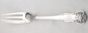 Een zilveren vork met gelobd vioolvormige steeluiteinde met smaller toelopende steel. Drietandige vork, de linker tand dient ook als snijmes. Uiteinde steel is gedecoreerd met schelpmotief en daaronder zit een gekroond WR monogram.