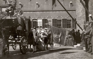Zwartwit foto van links de gouden koets waar Koningin Wilhelmina met prins Hendrik uitstapt. Rechts de trap naar de ridderzaal. Daarvoor soldaten met lansen en één met een standaard. Op de achtergrond meer soldaten. Er hangen mensen uit de ramen op de achtergrond.