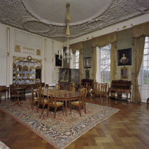 Vierkante foto van een kamer met een parketvloer en een stucwerkplafond met medaillons. Links een wand met een cremekleurige lambrizering met gouden ornamenten. In het midden bovenin twee deurtjes met daaronder een nep-balustrade. Daaronder een nis met spiegels en een goudkleurige klok erboven. In de nis staat blauw keramiek. Midden op de vloer ligt een tapijt met daarop een houten ovalen tafel met daaromheen acht stoelen. In de rechterwand zitten drie ramen, daartussen is de muur bespannen met groen textiel en er hangen schilderijen en spiegels. Er hangen geel/gouden gordijnen met lambrequins. Tussen de ramen staan commodes. In de hoek staat een beschilderd kamerscherm. In het midden hangt een eenvoudige kroonluchter.