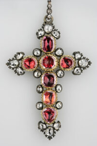 Een kruis bestaand uit zeven rood fonkelende robijnen, omgeven door twintig diamanten. Het geheel is gevat in goud en zilver.
