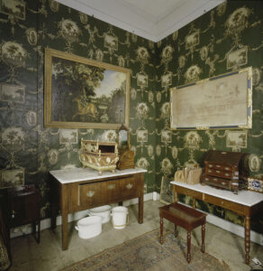 Foto van de hoek van een kamer. Op de muur een groen behang met daarop allemaal gedrukte medaillons en ingelijste prenten. Aan de linkerwand hangt een schilderij met een hert, aan de rechterkant een ingelijste stamboom. Onder elk schilderij staat een tafel met daarop kisten en kastjes. Bij de rechter tafel staat een kruk, onder de linker tafel staan keramieken bakken en een keramieken emmer. De vloer is van hout en we zien nog net een hoek van een versleten vloerkleed.