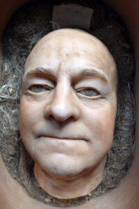 Een afdruk van het hoofd van een kale man, met dunnen lippen. Het hoofd ligt op grijze krullen.