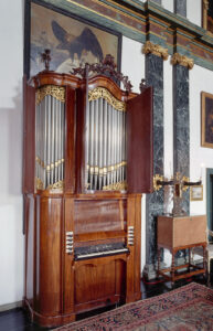In een hoge ruimte staat een orgeltje. Het heeft een mahoniehoutenkast die aan de zijden afgerond is. De deurtjes van de bovenkant staan open, waardoor je de pijpen zit. Eronder zit één klavier dat aan weerszijden vijf registerknoppen heeft. De omgeving is rijk aangekleed: boven het orgel hangt een schilderij van een adelaar, op de zwarte vloer ligt tapijt en tegen de witte wanden zitten donker marmeren pilasters met daarop lichtarmaturen van zwarte onderarmen die een kandelaar vasthouden.