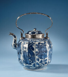 Theepot van porselein met een geribd, bolvormig lichaam en S-vormige tuit, beschilderd in onderglazuur blauw. De wand is verdeeld in vakken met afwisselend een bloeiende plant (chrysant, pioen) en een persoon zittend op een omheind terras; de schouder met een band servetwerk afgewisseld met een bloem in een cartouche. Voorzien van een 19e-eeuws zilveren montuur en handvat door Arnoldi & Wielick, Amsterdam 1850. Kraakporselein.