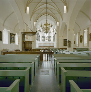 Interieur van een kerk richting het koor. Op de voorgrond groene kerkbanken. In het midden een preekstoel met daaromheen een bruin hek. Rechts tegenover de preekstoel eveneens groene banken. Aan de wanden en rondom in het koor hangen rouwborden.